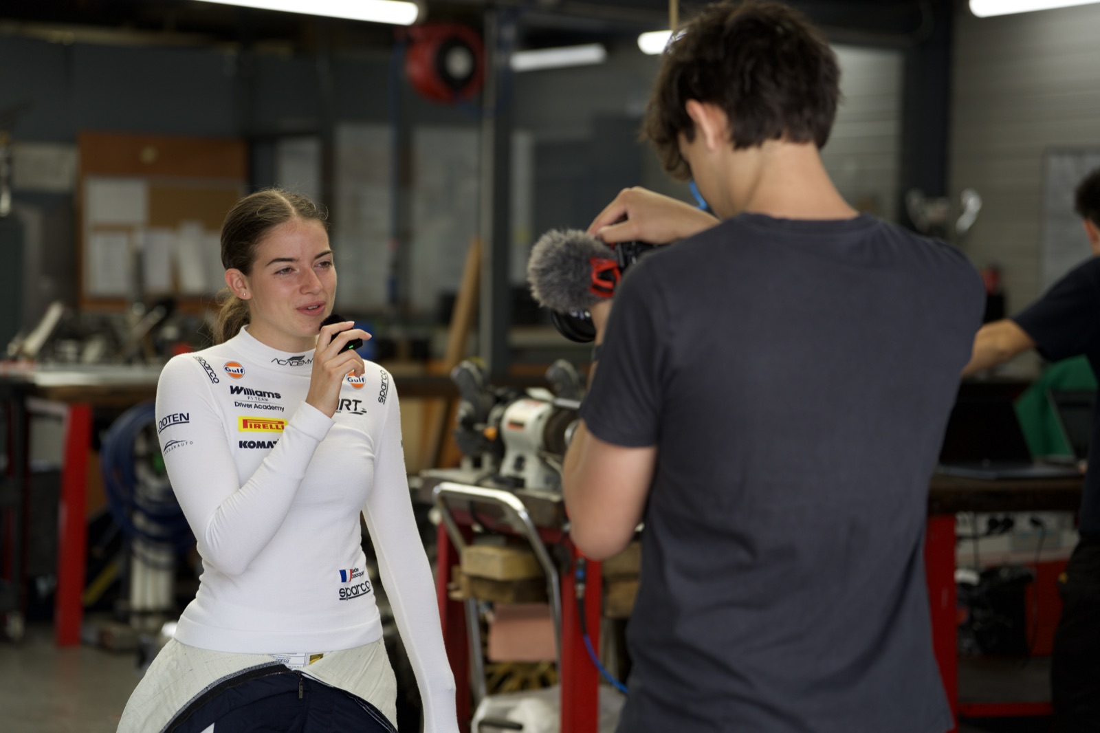 Lucas caméra à l'épaule en interview d'une pilote automobile dans un atelier